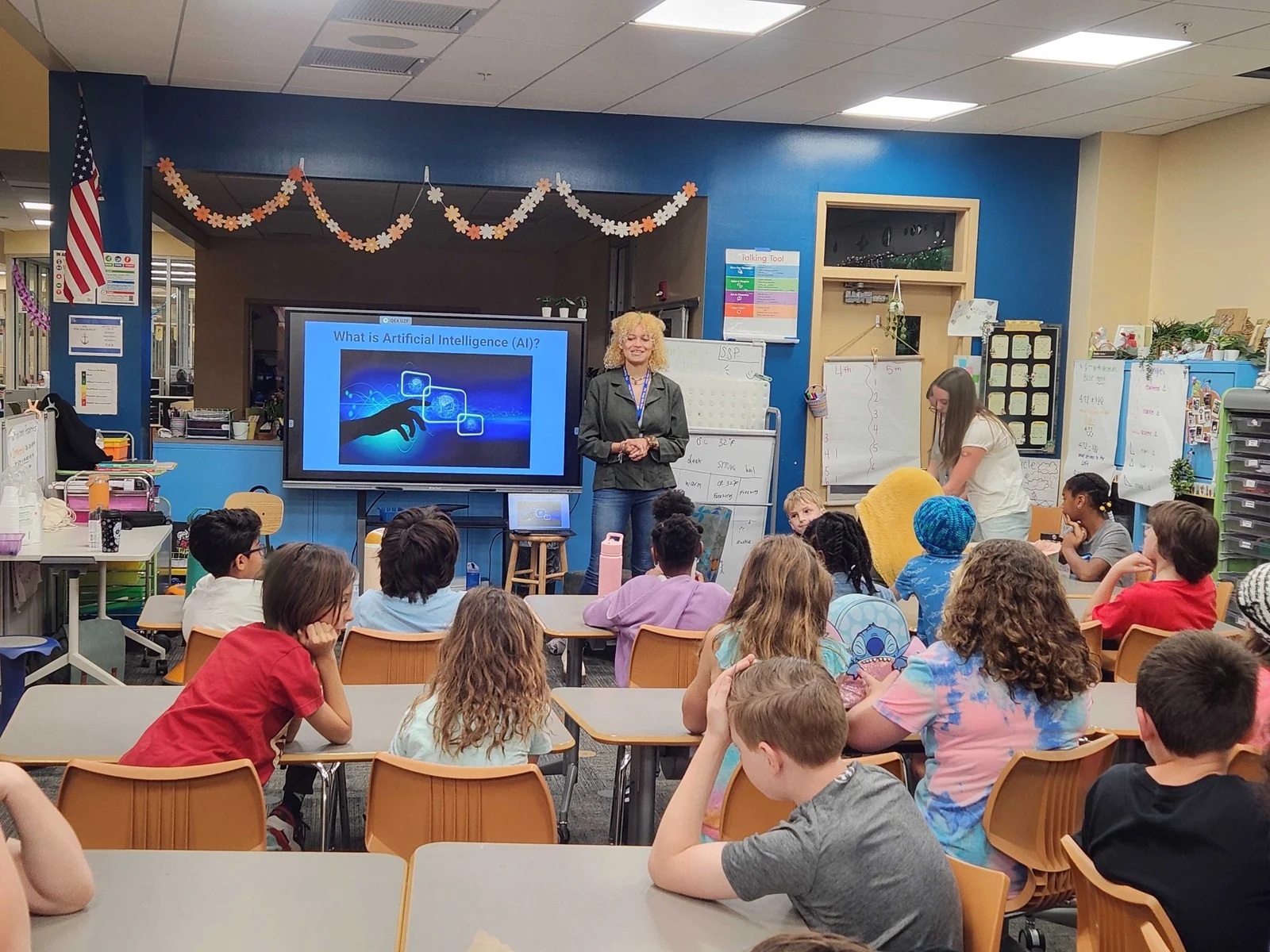 A teacher addresses a middle school classroom while standing next to a monitor displaying the text "What is Artificial Intelligence (AI)?"
