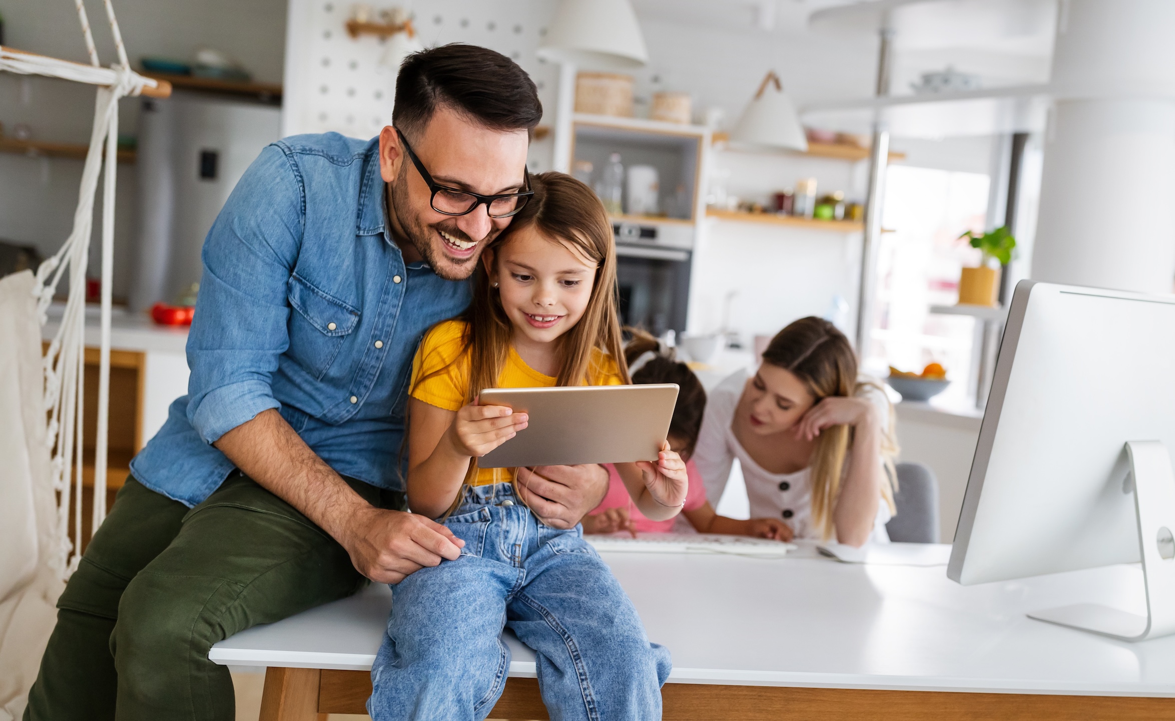 Father and daughter looking at a tablet, with two other children in the background working at a table.