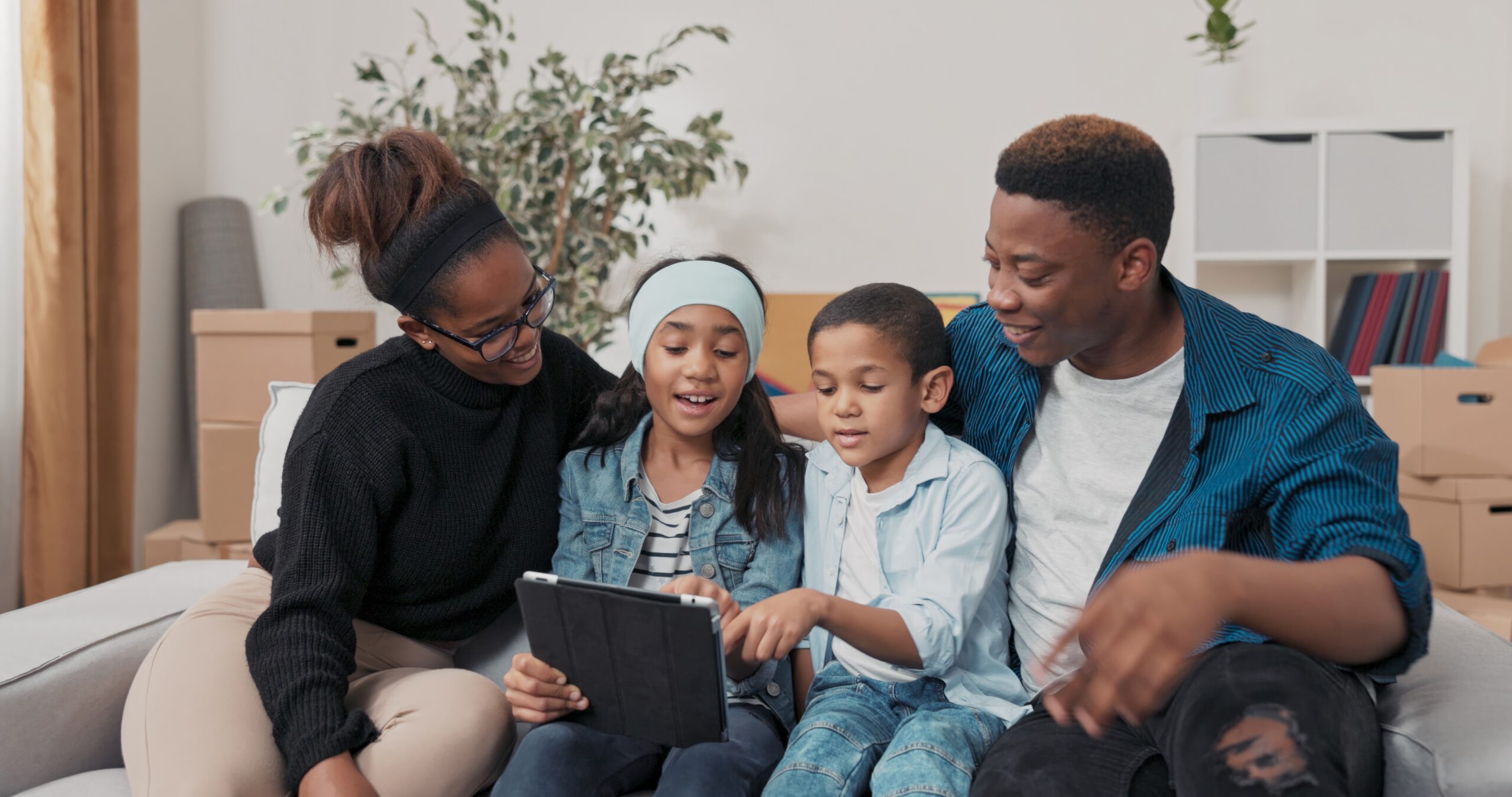 Family of four looking at a tablet and smiling on a white couch in a living room.
