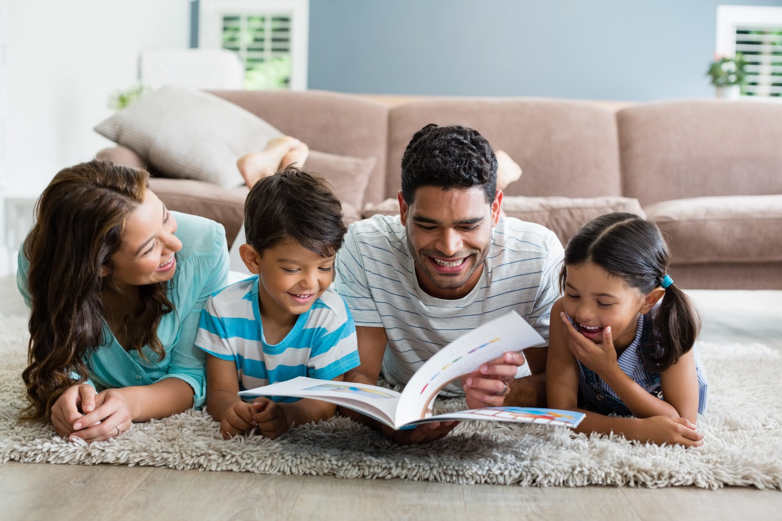 Family of four lying on the floor reading a book together in a living room.