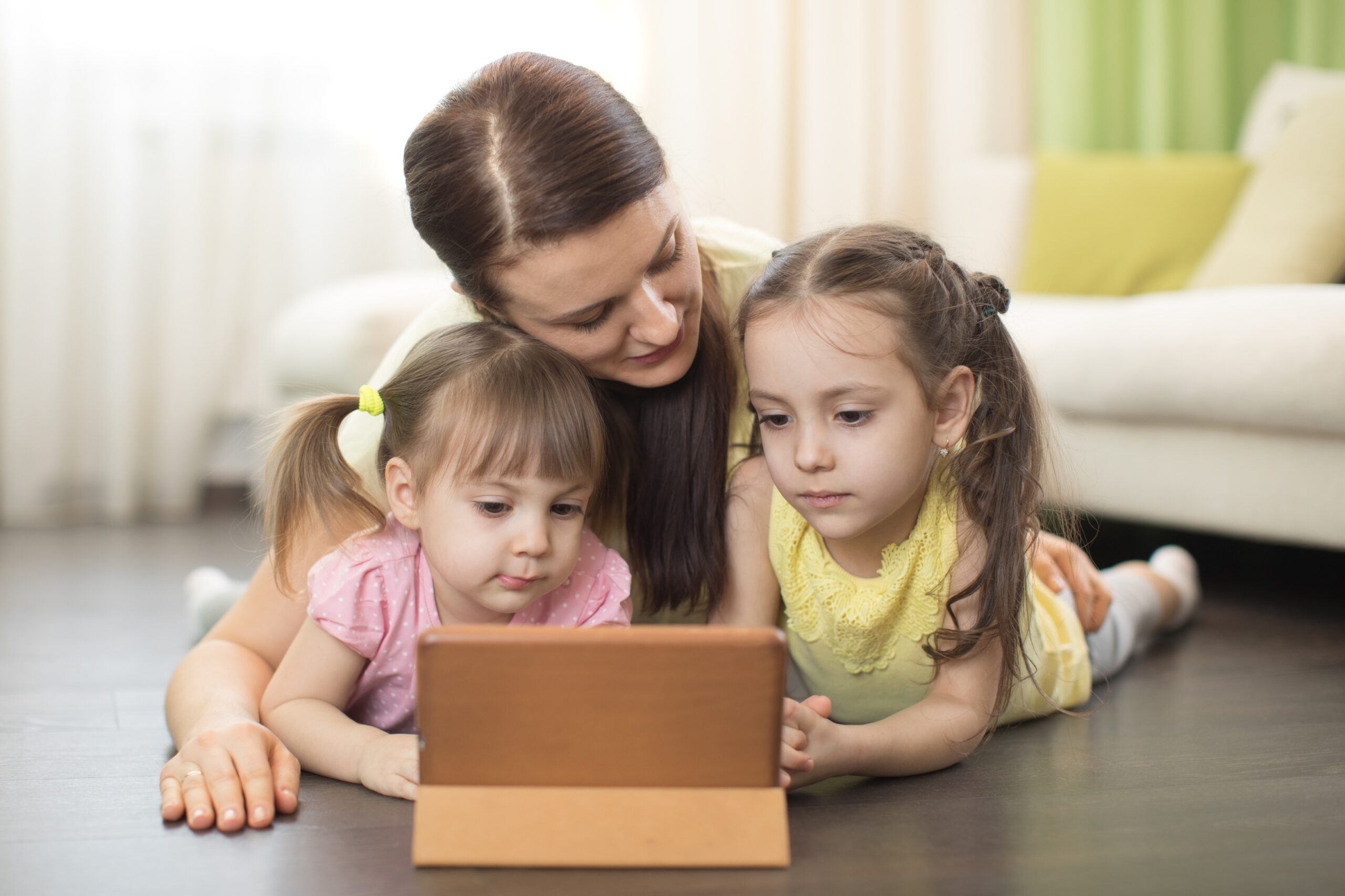 Mother and two young daughters lying on the floor, looking at a tablet.
