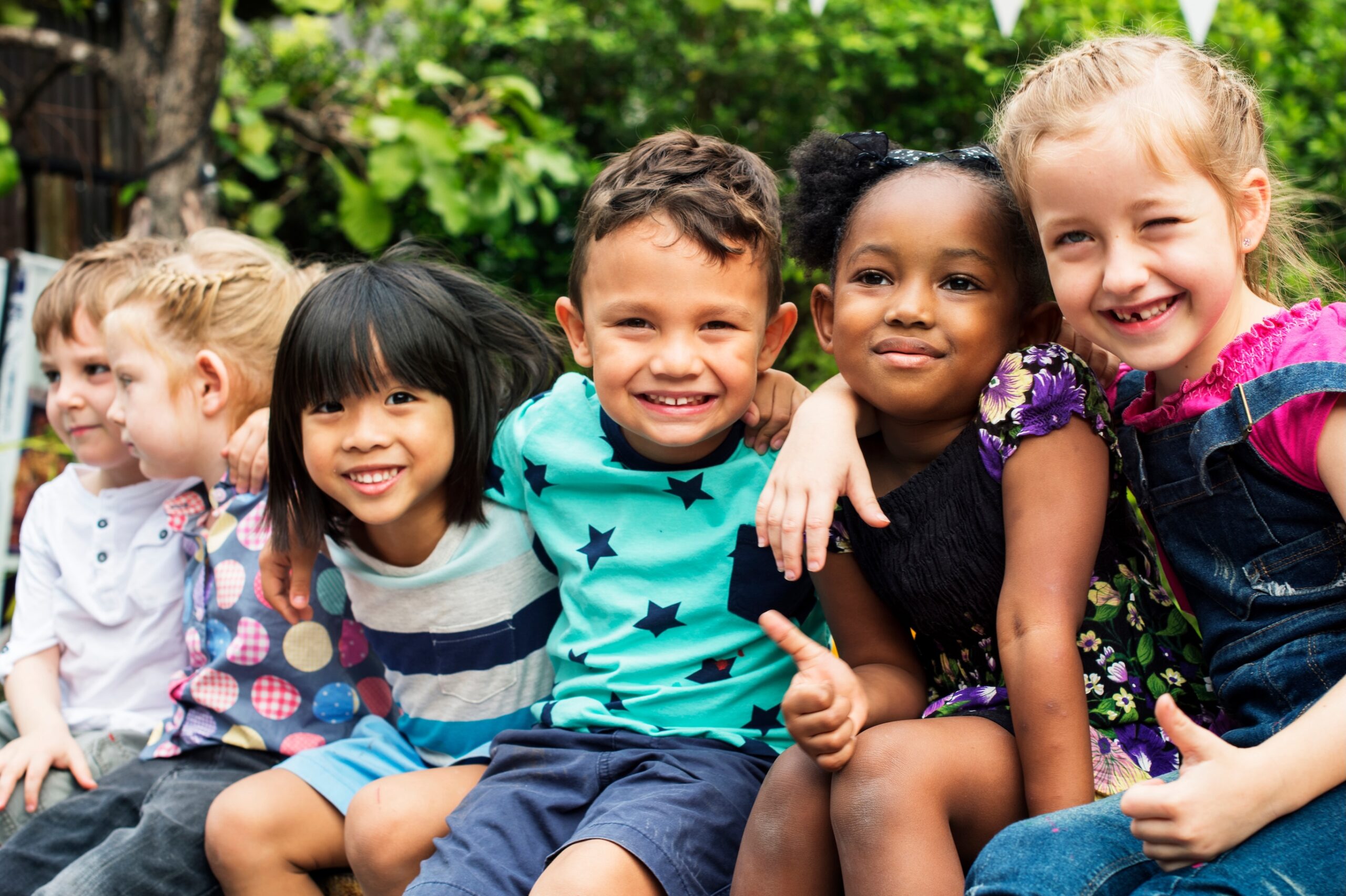 Five smiling children of diverse ethnicities pose closely together outdoors.